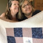 Paola Jo and Quilter smiling for camera as they hold up a blue and pink quilt